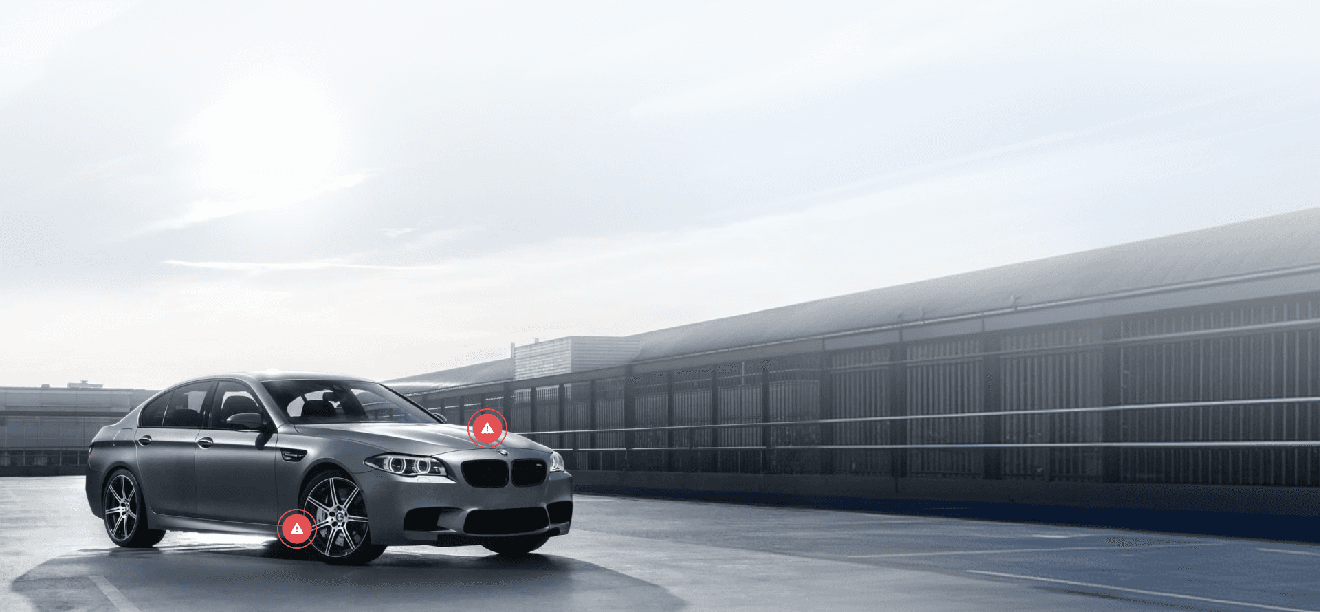 Una BMW argento parcheggiata su una superficie di cemento, con due icone rosse di avviso con punto esclamativo: una sul cofano e l'altra vicino alla ruota anteriore destra.